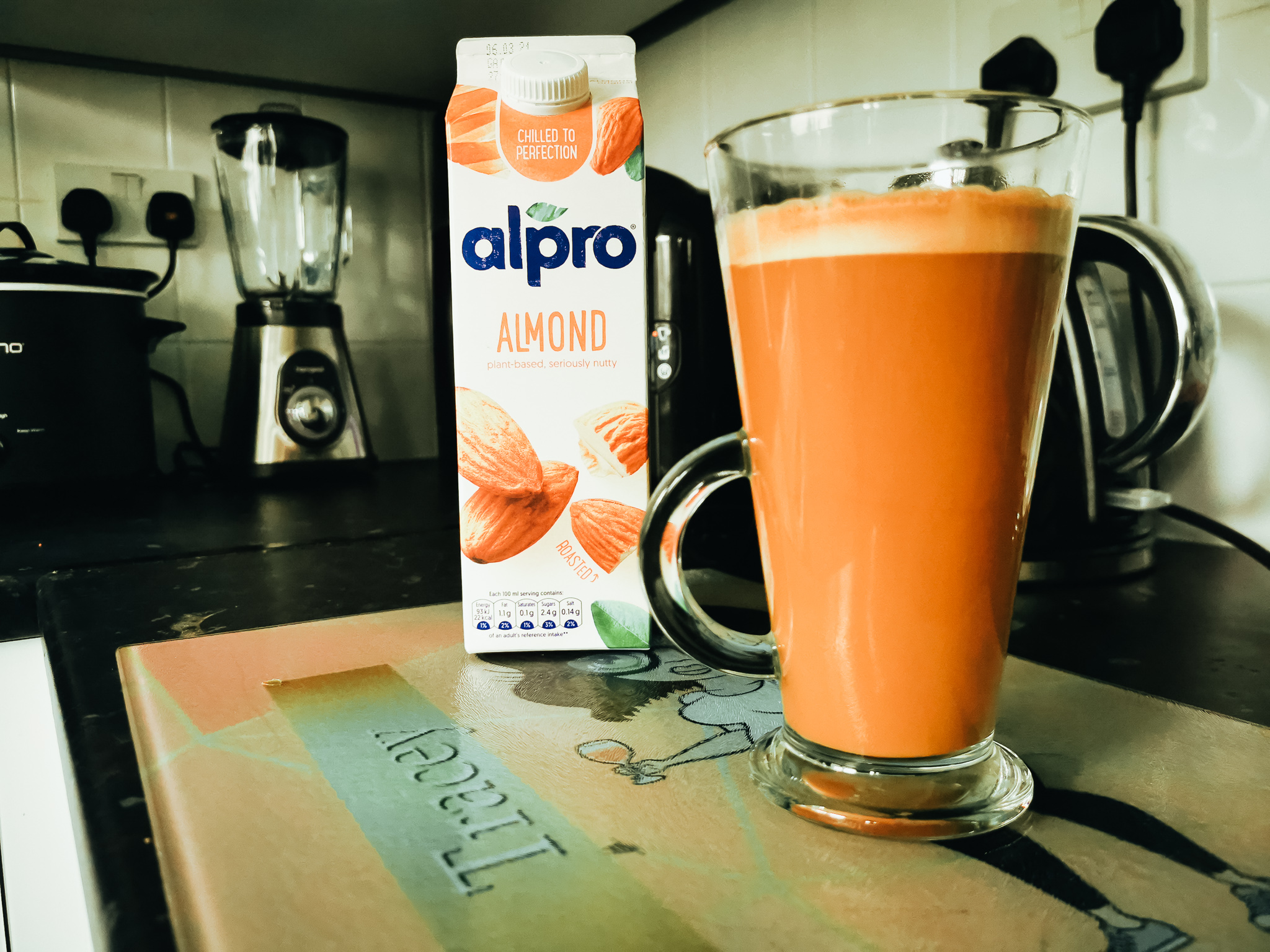 close up of a latte in a clear glass tall cup in front of a carton of alpro almond milk surrounded by kitchen appliances