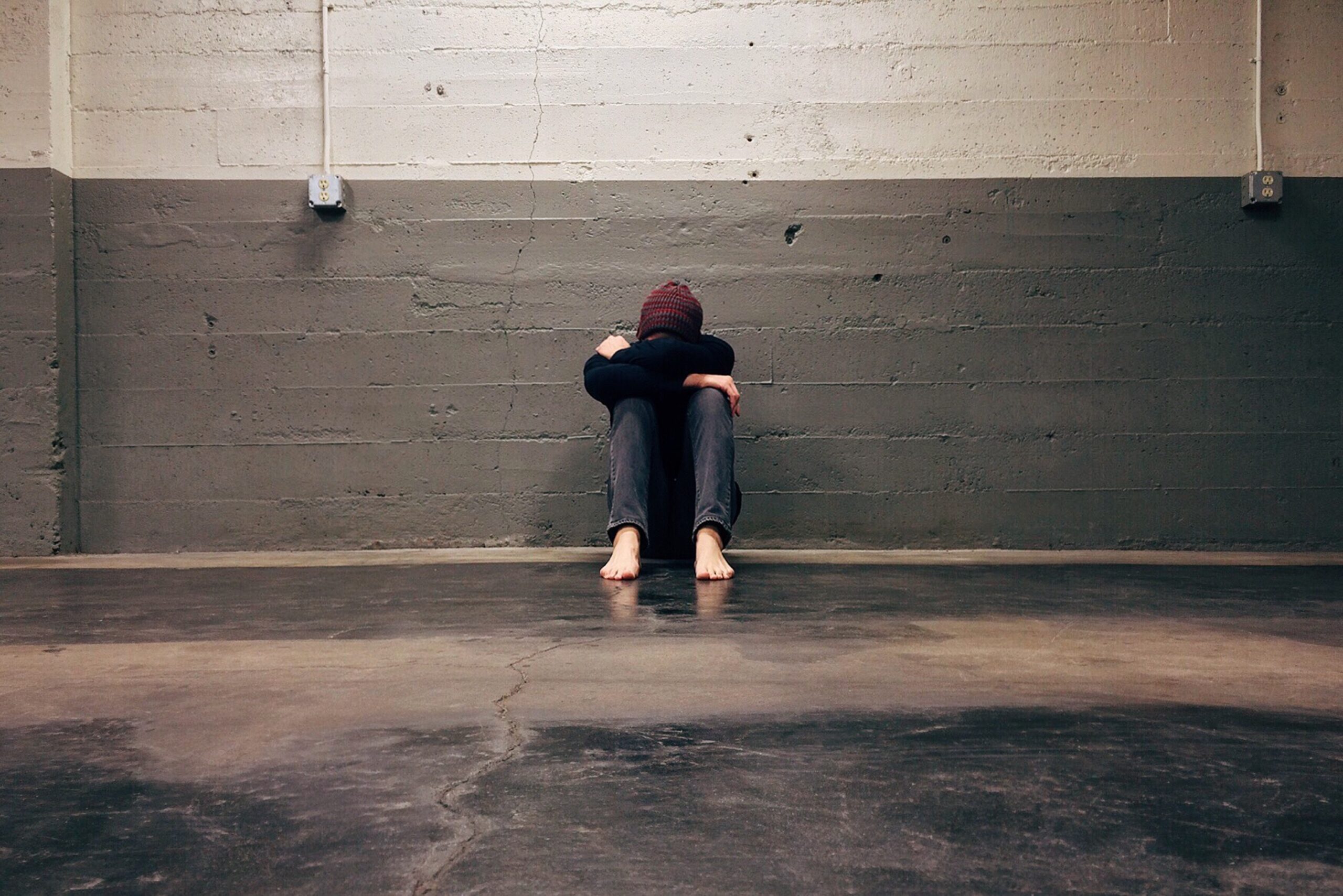 person sitting in front of a grey and white wall with their head on thier knees in a sitting fetal position