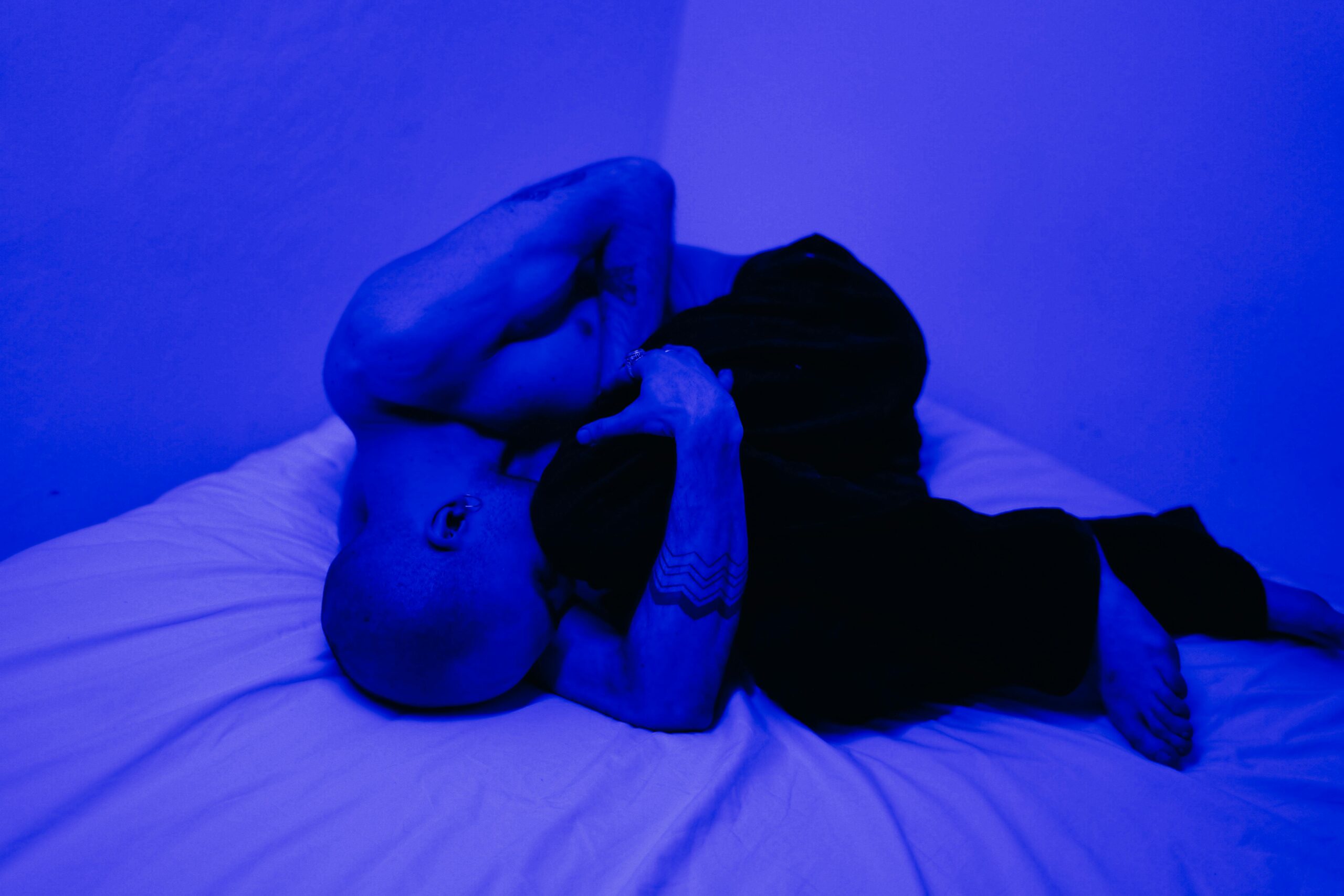 a blue filtered image of a man curled up on a bed imitating being in pain